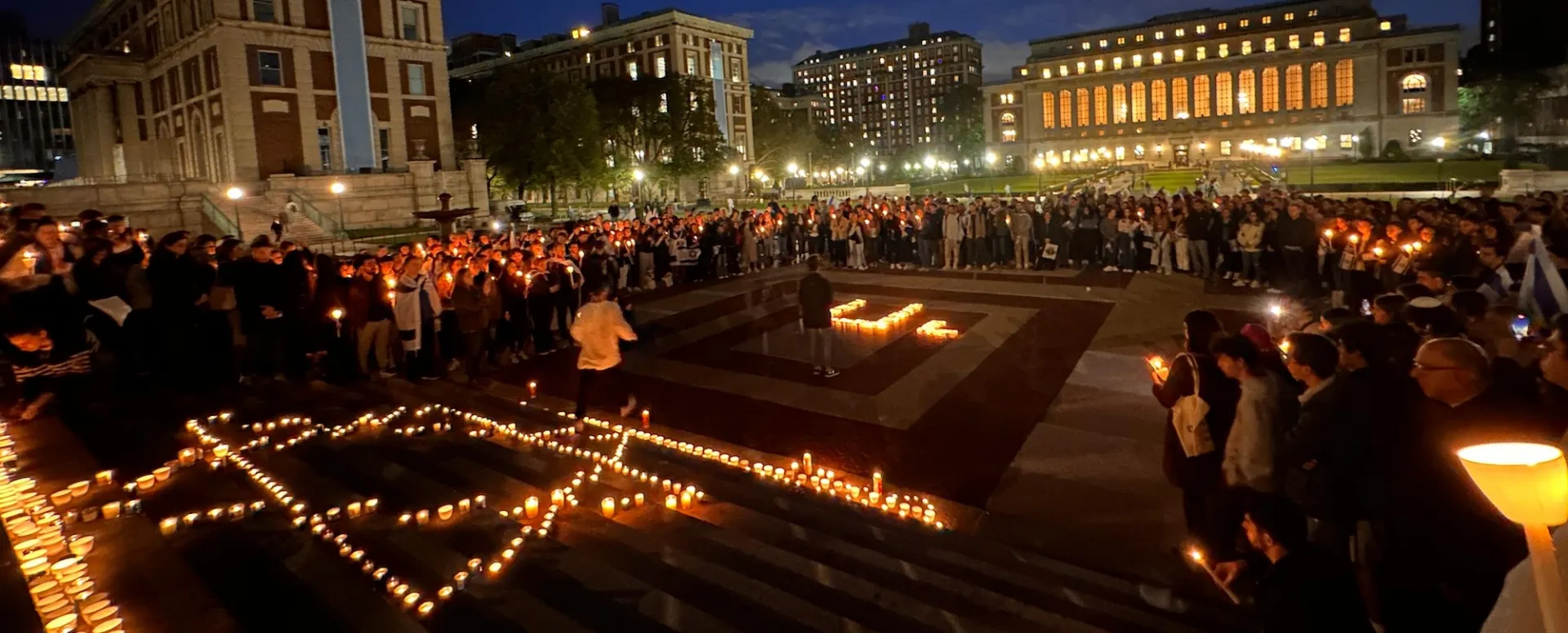 Countless people holding lit candles outside the Low library steps at night. On the ground, lit candles make the shape of star of David and "chai" in Hebrew letters